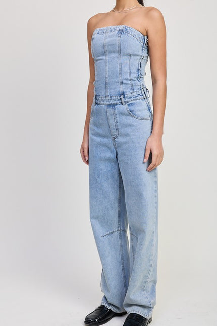 A full-body view of a model wearing a strapless denim jumpsuit in light-wash blue. The jumpsuit features a fitted bodice with vertical seam details, a side zipper, a belted waist, and wide-leg jeans with subtle stitching accents. 